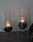 Two clear glass candle holders with candles and decorative elements on a neutral background