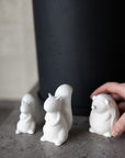 Three white ceramic squirrel figurines on a dark surface with a hand reaching towards one of them.