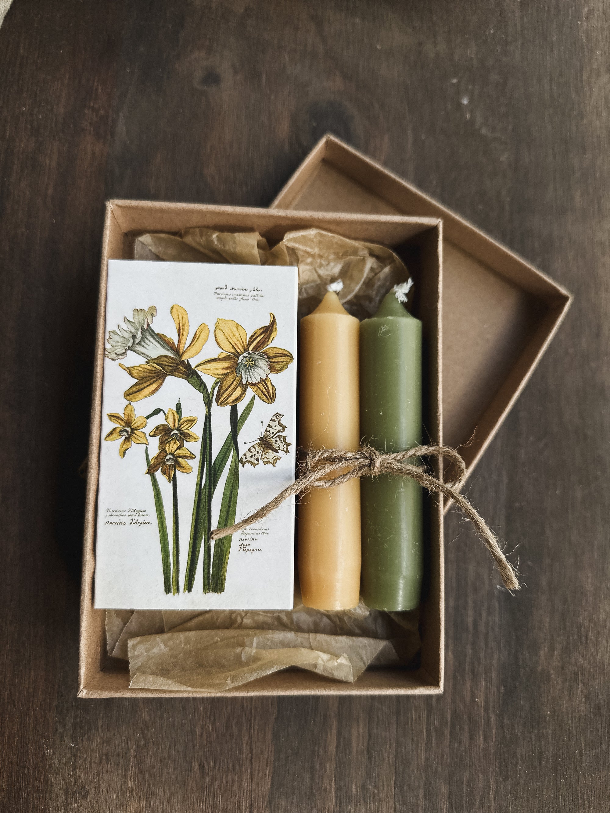 A gift box containing a vintage daffodil illustration and two candles in matching colors, tied with a string.
