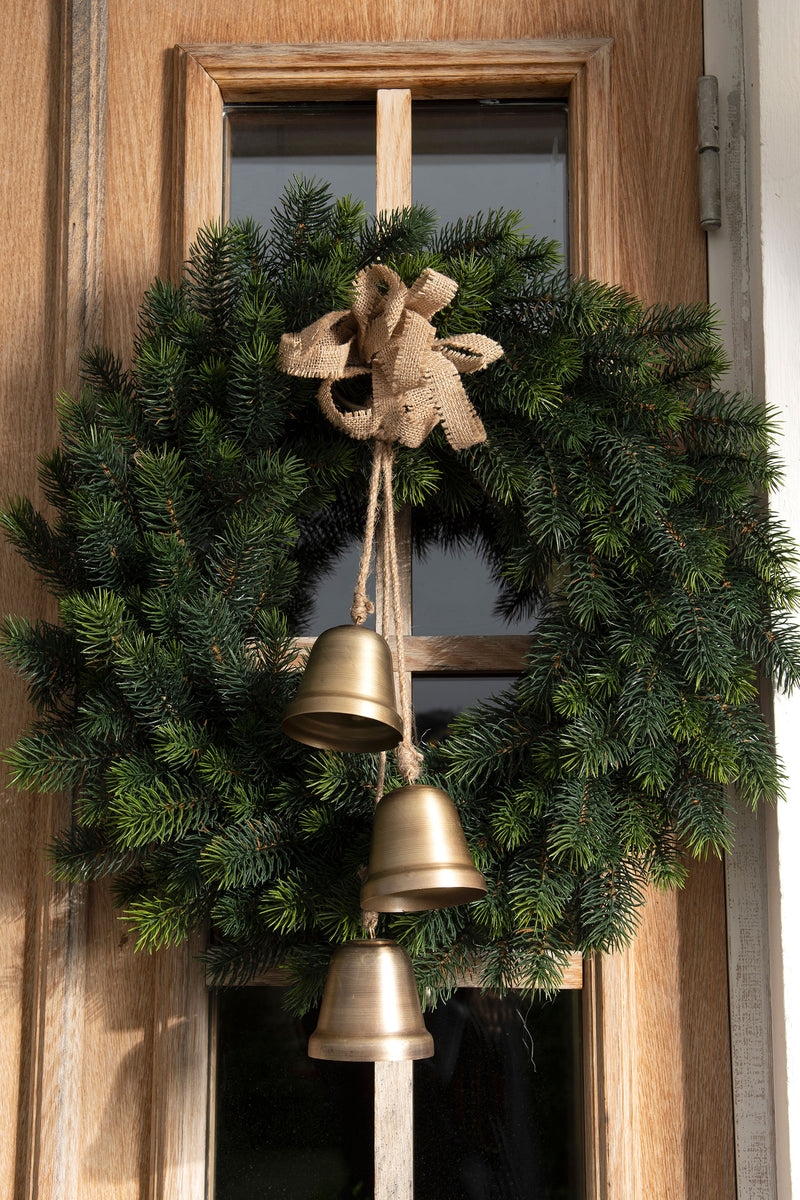 Green wreath with gold bells and a burlap bow hanging on a wooden door.