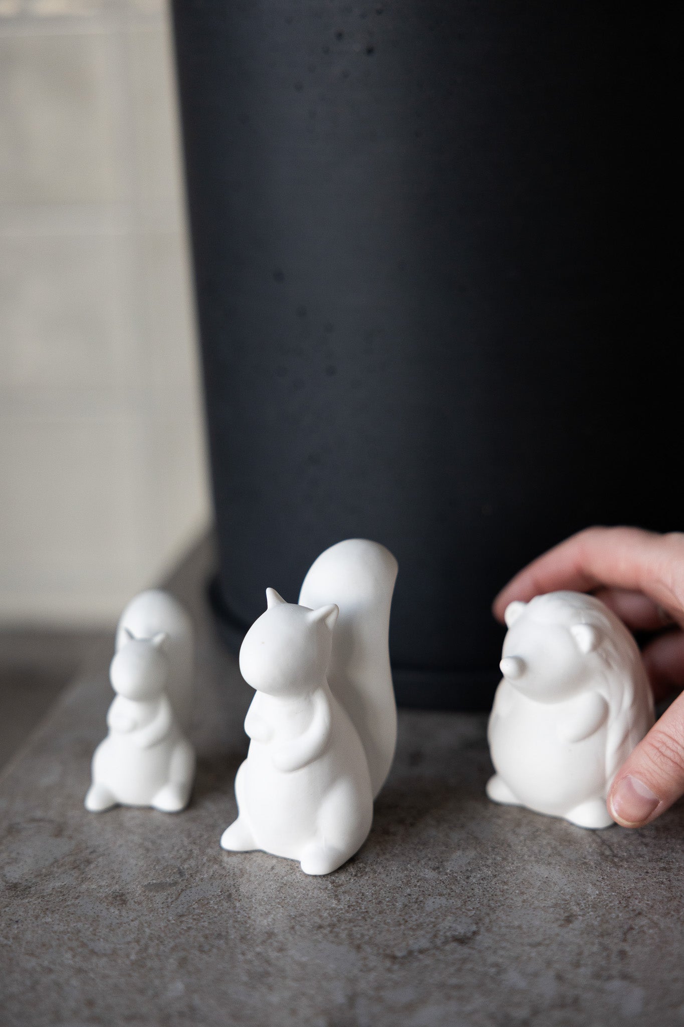 Three white ceramic squirrel figurines on a dark surface with a hand reaching towards one of them.