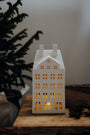 White house-shaped lantern with lights on a wooden surface next to a plant