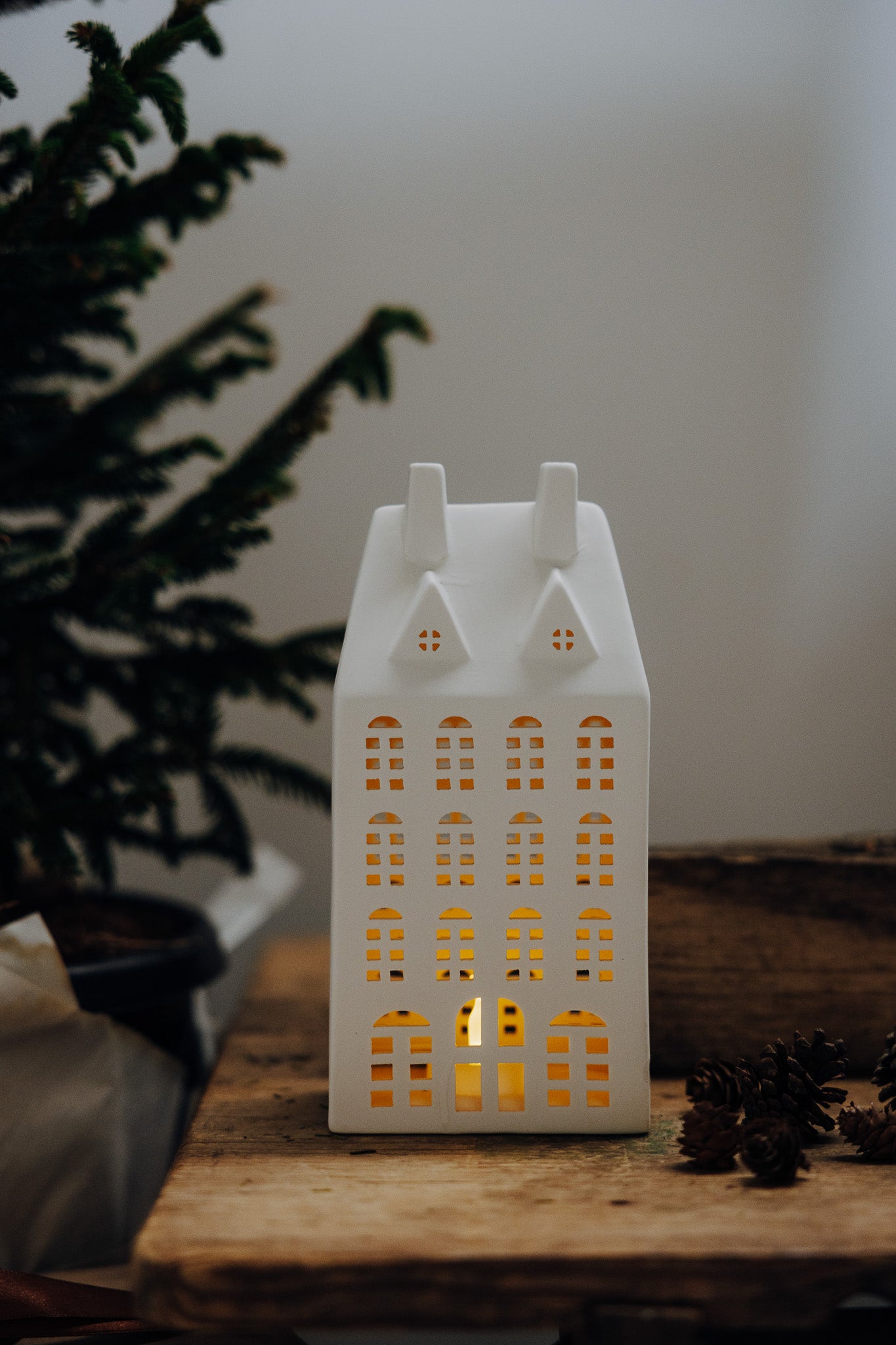 White house-shaped lantern with a warm glow on a wooden surface, surrounded by pine branches.