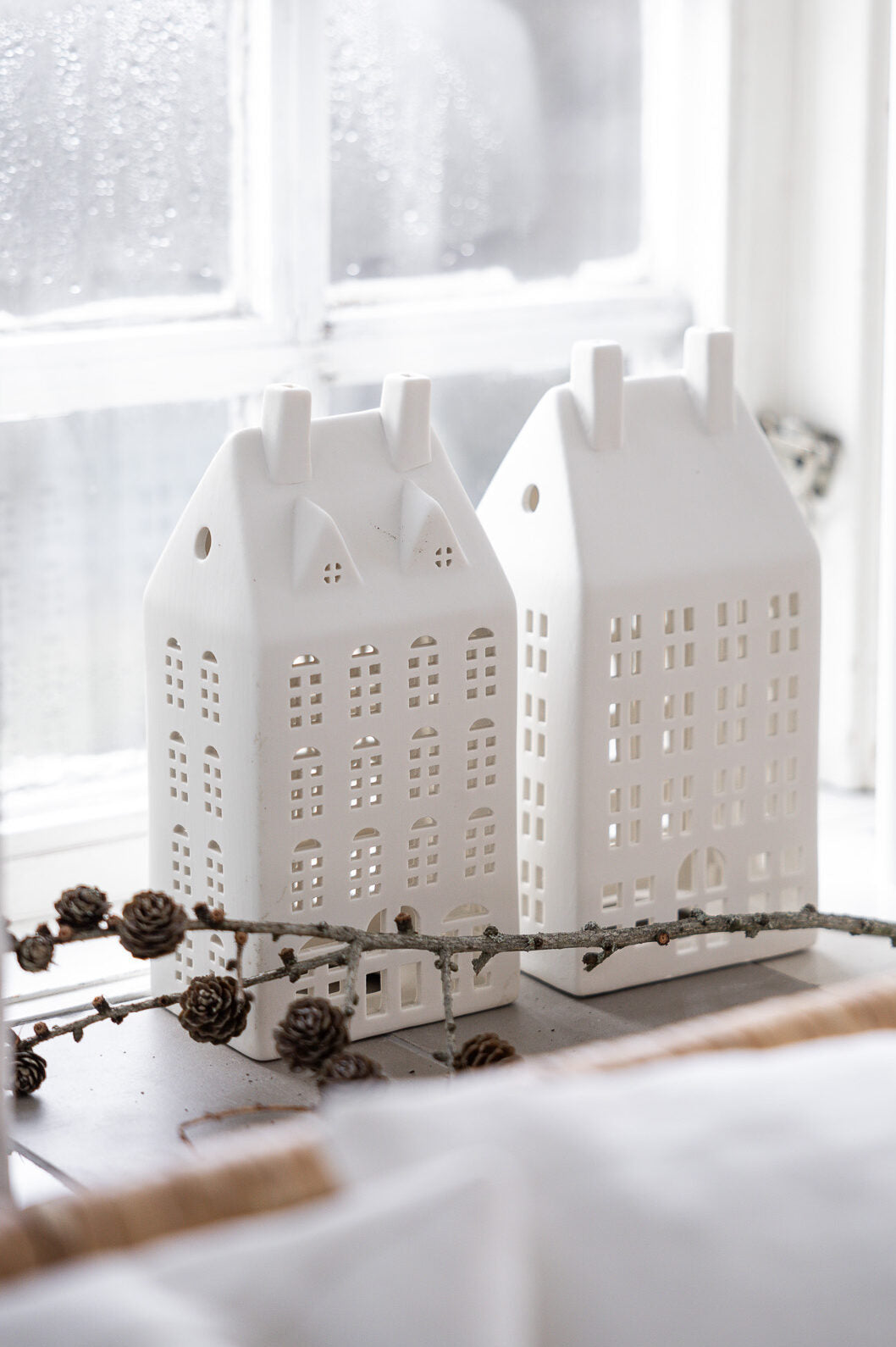 Two white ceramic house-shaped lanterns on a windowsill with a snowy background.