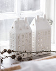 Two white ceramic house-shaped lanterns on a windowsill with a snowy background.
