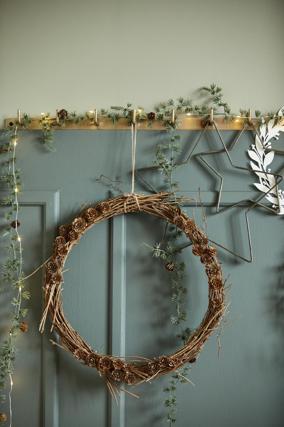 Decorative wreath with pinecones and greenery on a teal wall with a gold candle holder and star decorations.